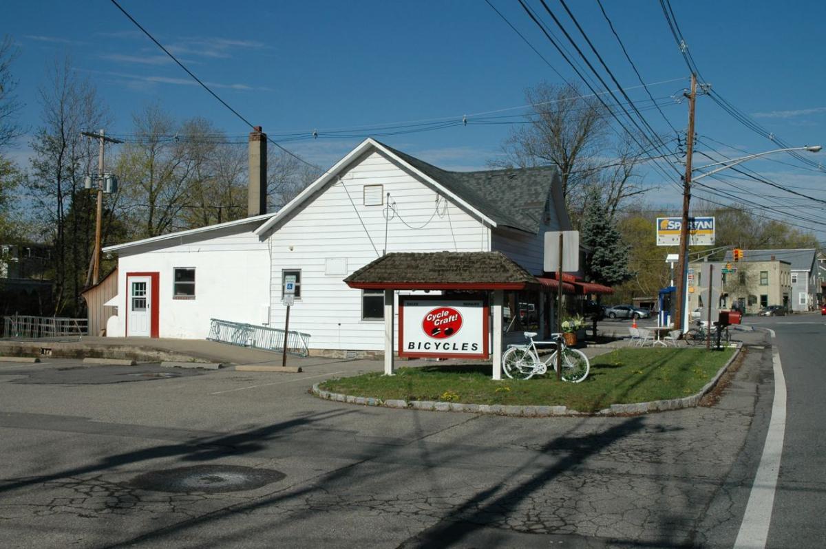 A corner bicycle shop with a white exterior and a red roof sign that reads "Cycle Craft! Bicycles." A white bicycle is displayed near the entrance, and there are power lines overhead. The surrounding area features a paved parking lot and grassy patches.