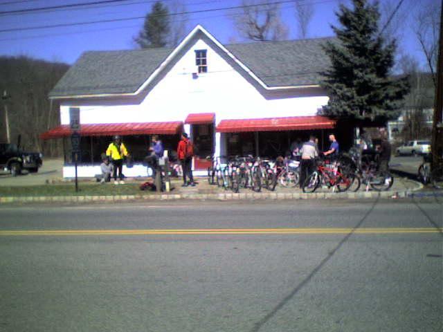 A white building with a gray roof and red awnings, featuring a small group of people gathered outside. Several bicycles are parked along the sidewalk. The scene is set on a sunny day with a clear blue sky and trees in the background.