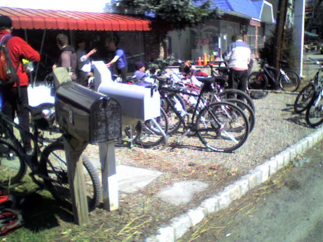 A group of people gathered outside a building with a red awning, with several bicycles parked nearby. In the foreground, there is a mailbox next to a bike. The scene suggests a community or social gathering, possibly related to cycling.