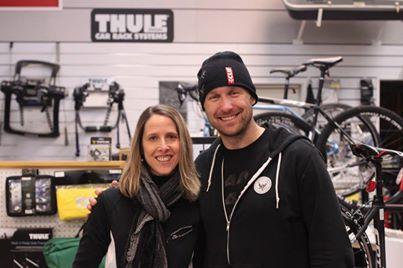 A man and a woman smile together for a photo in a store that features bike racks and accessories displayed on the walls behind them. The woman has shoulder-length hair and wears a black jacket, while the man is wearing a black beanie and hoodie.