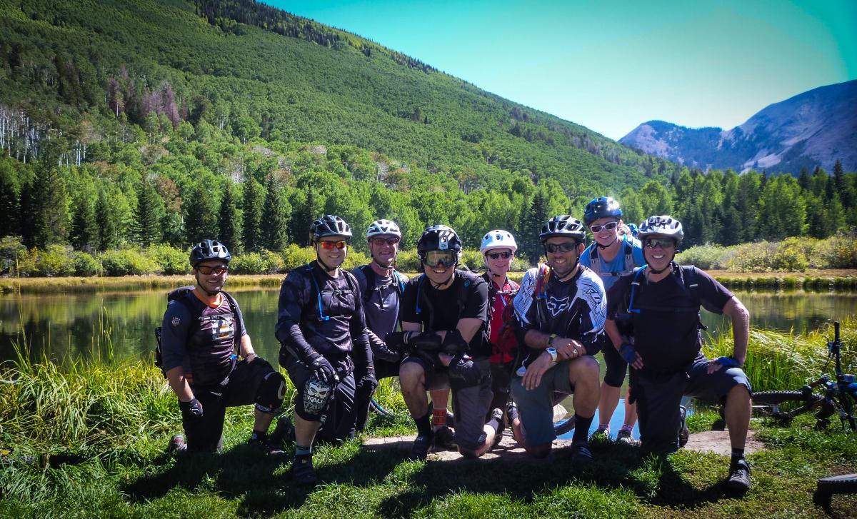A group of eight mountain bikers posing together by a serene lake, surrounded by lush green forests and mountains in the background. They are wearing helmets and biking gear, smiling at the camera. Porcupine Rim mountain bike trail.