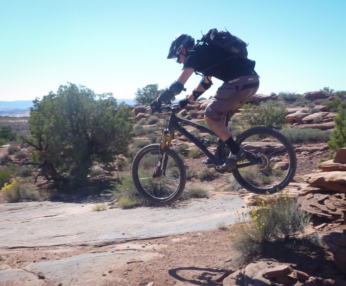 A mountain biker in a helmet and protective gear is mid-jump over rocky terrain, showcasing an action shot of mountain biking in a dry, mountainous landscape with sparse vegetation. Porcupine Rim mountain bike trail.