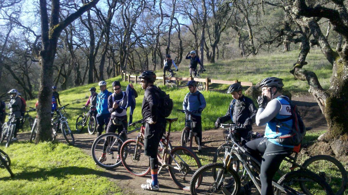 A group of mountain bikers gathered on a dirt path in a wooded area, surrounded by trees. Some riders are adjusting their gear or preparing for a ride, while others are standing next to their bikes. In the background, a few riders are on an elevated wooden platform, with green grass and trees creating a natural setting.