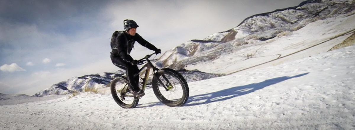 A person riding a fat tire bike on a snowy trail, surrounded by mountains. The cyclist is dressed in a black outfit and a helmet, navigating the snowy landscape under a partly cloudy sky. North Table Mountain mountain bike trail.
