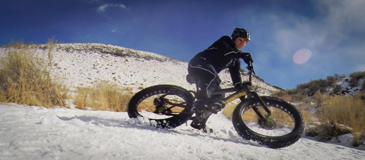 A person riding a fat tire bike on a snowy trail, surrounded by a snowy landscape and sparse vegetation, with a blue sky above and a few clouds in the background. North Table Mountain mountain bike trail.