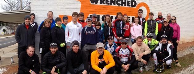A group of cyclists and supporters pose for a photo in front of a vibrant mural that says "WRENCHED." The group, consisting of both men and women of various ages, is dressed in cycling gear and casual clothing, smiling and standing together outdoors.
