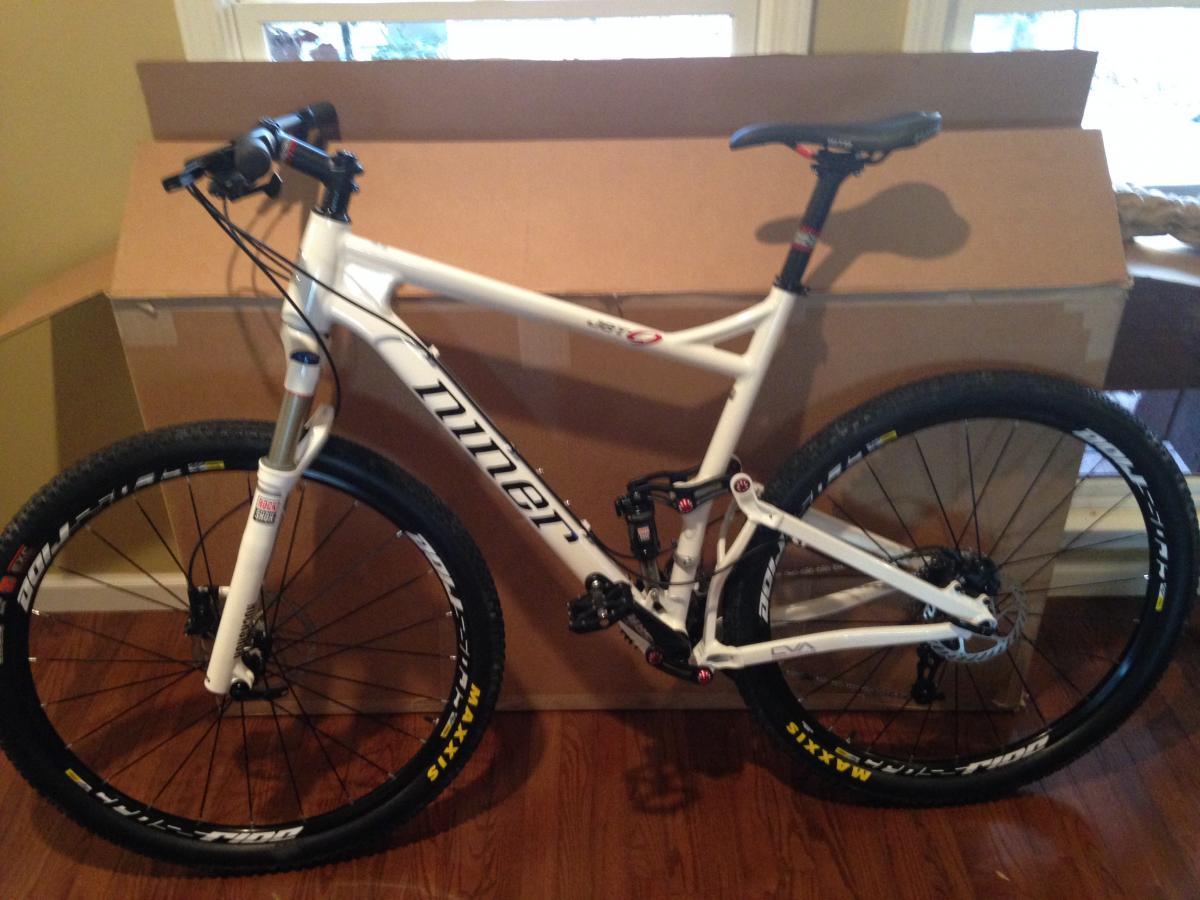 Niner Jet 9: A white mountain bike with black wheels and visible branding, positioned next to a large cardboard box in a room with hardwood flooring and windows in the background.