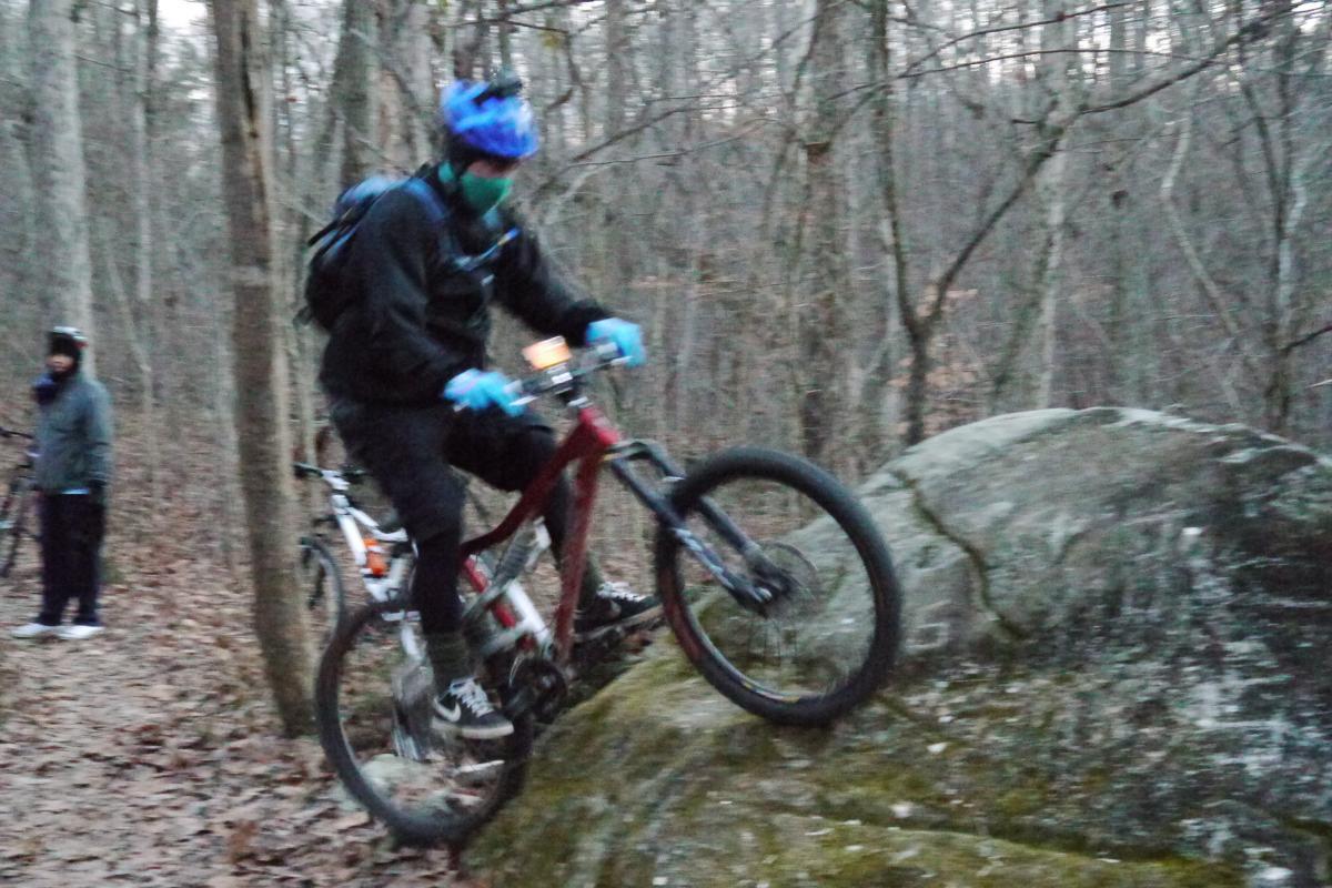 A mountain biker skillfully rides over a large rock in a wooded area during dusk, with another cyclist visible in the background. The rider is wearing a blue helmet, mask, and gloves, while the surrounding trees are bare, indicating it is likely autumn or winter. Hobby Park mountain bike trail.