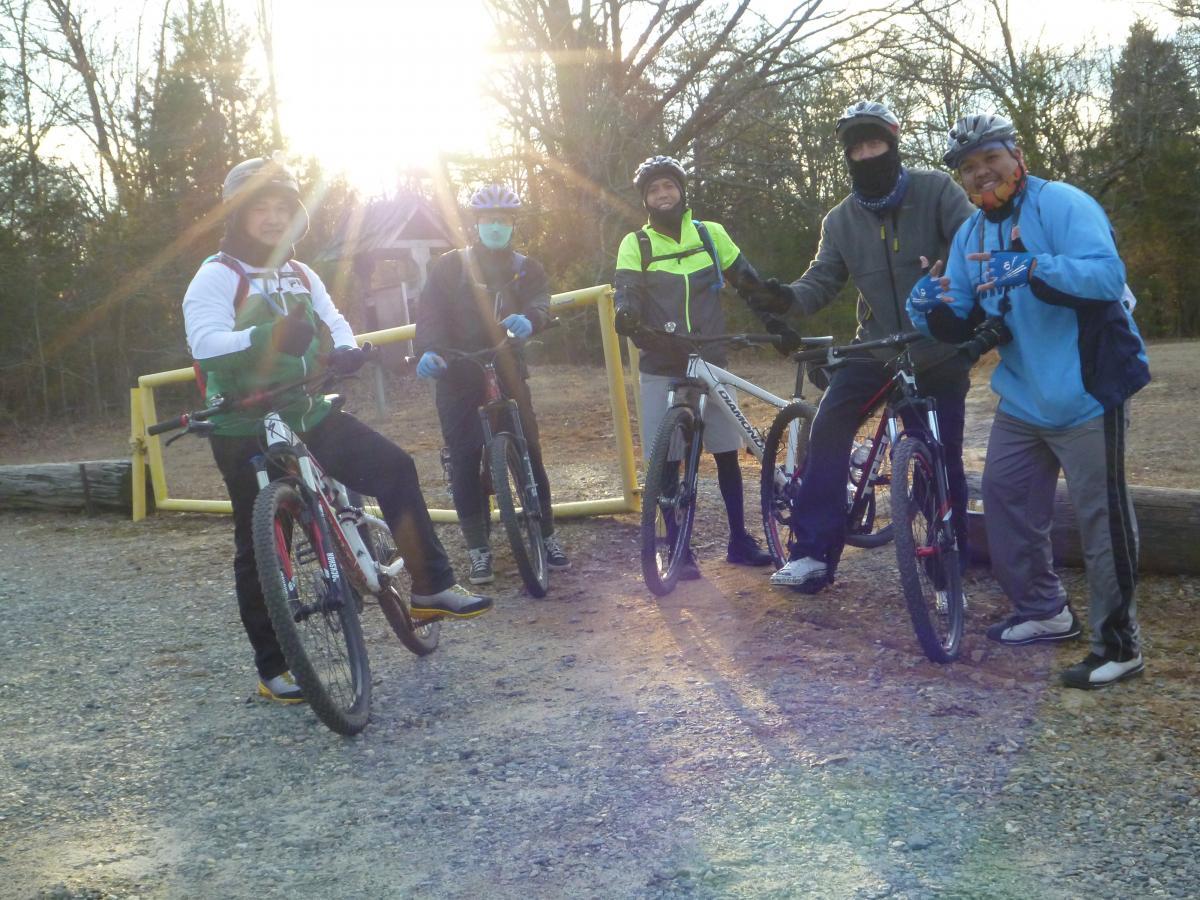 A group of five individuals wearing biking gear stands next to their bicycles in a wooded area during sunset. They are posing and smiling at the camera, with some wearing helmets and gloves. The background features trees and a yellow gate. Hobby Park mountain bike trail.
