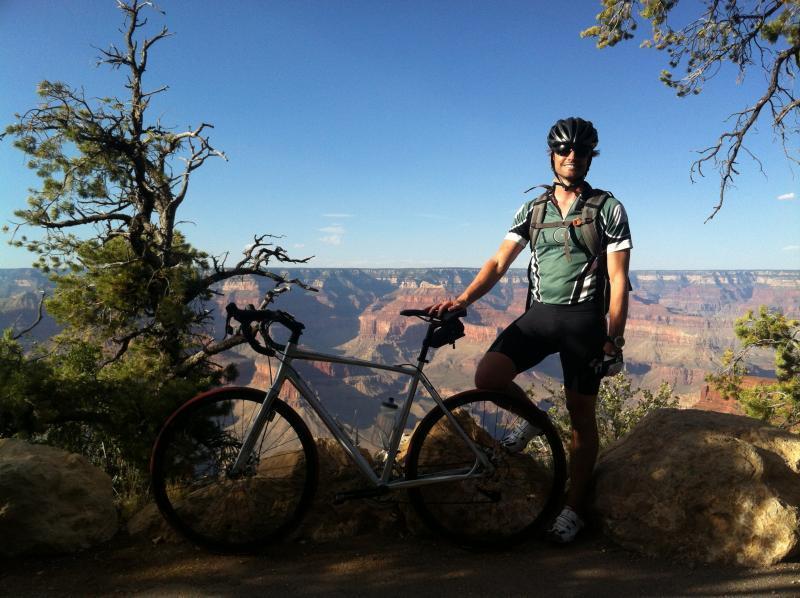 A cyclist wearing a green and black jersey stands next to a bicycle at the Grand Canyon, with scenic views of the canyon and blue sky in the background. Sedona, Arizona to Grand Canyon Road Cycling Tour mountain bike trail.