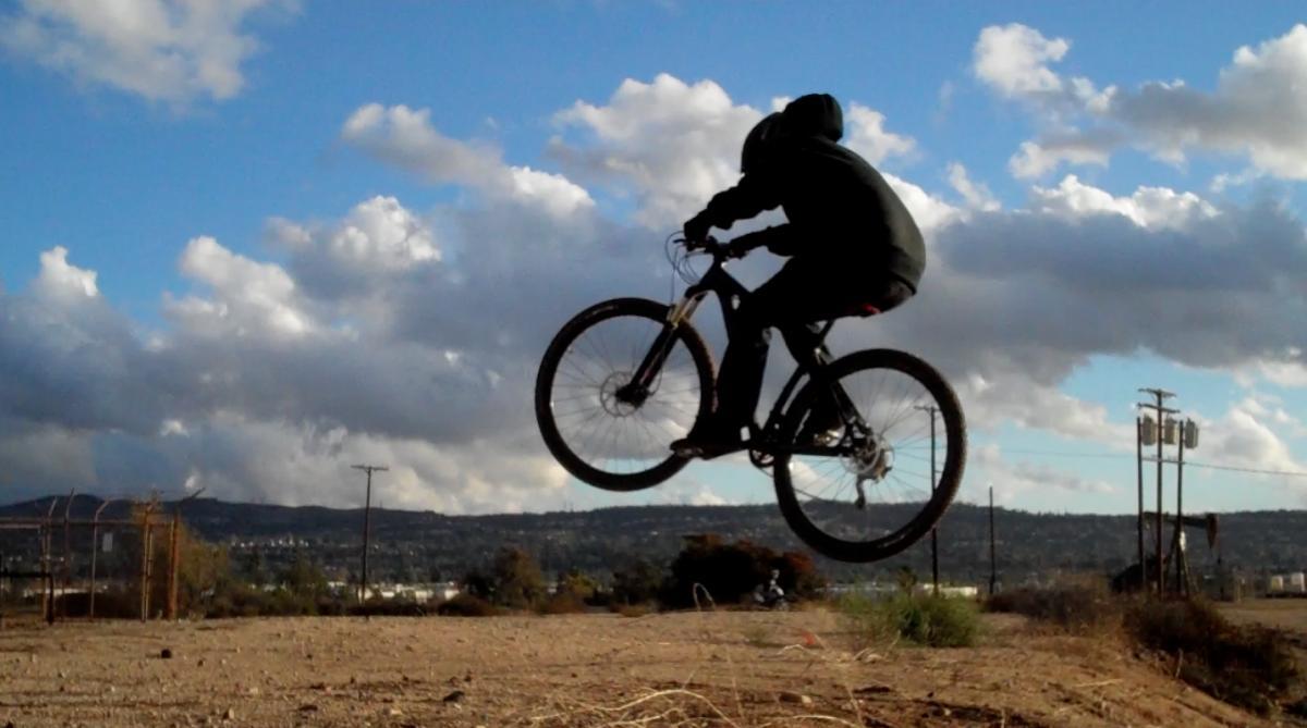 Specialized Crave Expert 29: A cyclist wearing a black hoodie performs a jump on a mountain bike over a dirt surface, with a backdrop of cloudy skies and distant hills.
