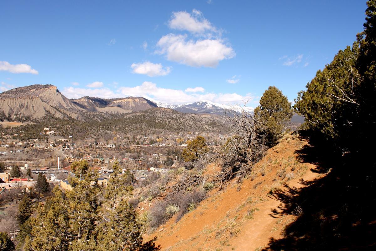 A scenic view of a mountainous landscape under a clear blue sky, featuring rocky hills and snow-capped peaks in the background. In the foreground, there is a winding dirt path bordered by scattered trees, leading down towards a small town nestled in the valley below. The town is visible among the greenery, with a mix of buildings and natural features. Horse Gulch mountain bike trail.