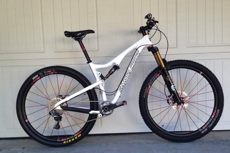 Santa Cruz Tallboy Carbon: A modern white mountain bike with black and red accents, featuring a full suspension frame, wide tires, and disc brakes, leaning against a garage door.