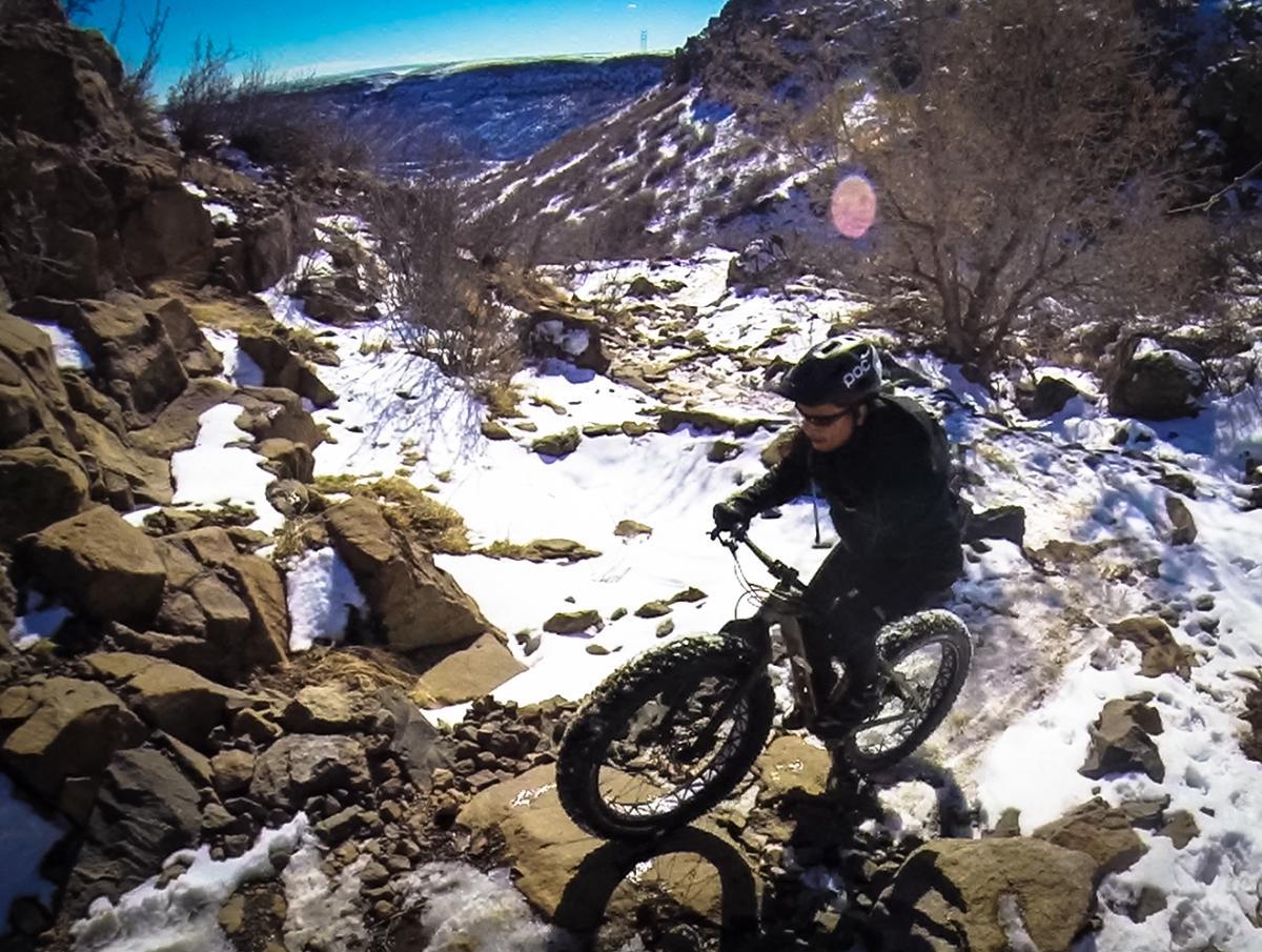 A person riding a fat tire bike on a rocky, snow-covered trail, surrounded by trees and mountains in the background. The scene captures the excitement of mountain biking in a winter landscape. North Table Mountain mountain bike trail.