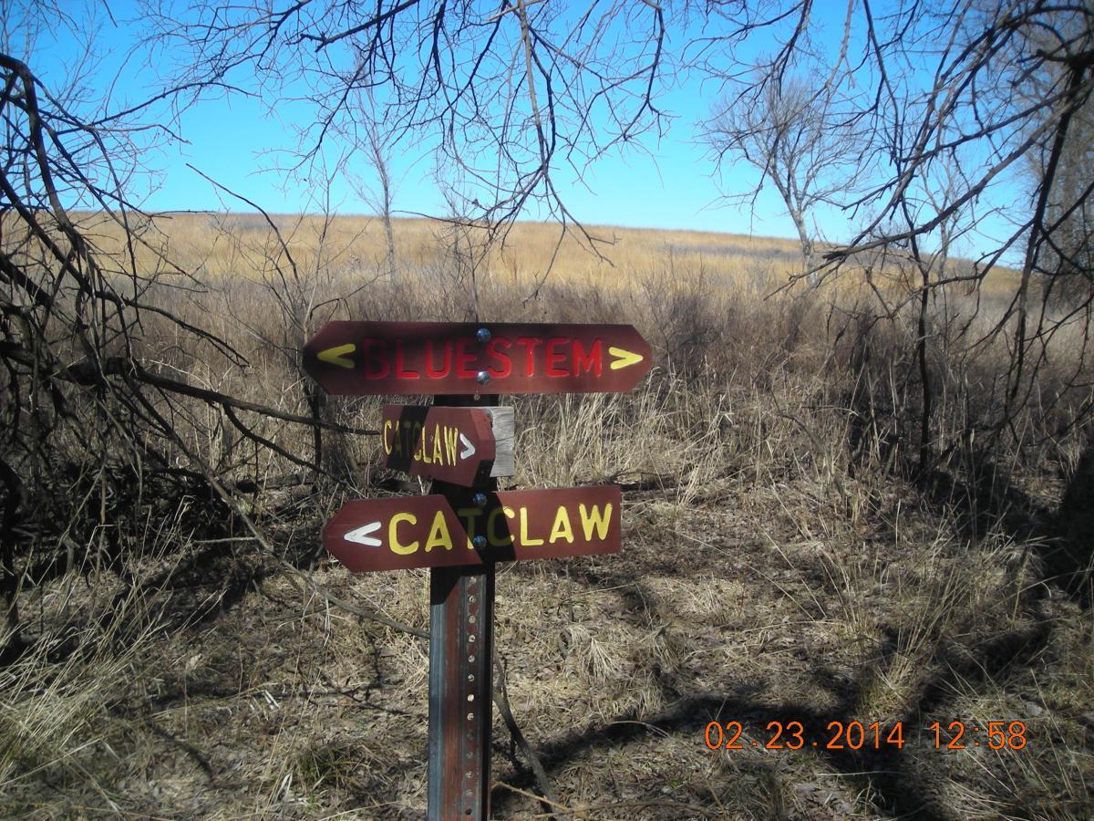 A wooden directional sign in a natural setting, indicating two trails: "Bluestem" and "Catclaw." The signposts are painted in dark brown with yellow arrows and text. Surrounding vegetation includes sparse grasses and bare branches, with an open field visible in the background under a clear blue sky. Fall River State Park mountain bike trail.