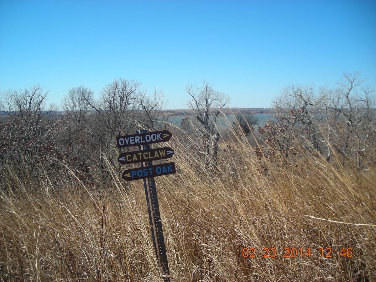 A directional sign with three arrows pointing to "Overlook," "Catclaw," and "Post Oak," set against a backdrop of tall grass and bare trees. The sky is clear and blue, suggesting a sunny day. The landscape includes a body of water visible in the distance. Fall River State Park mountain bike trail.