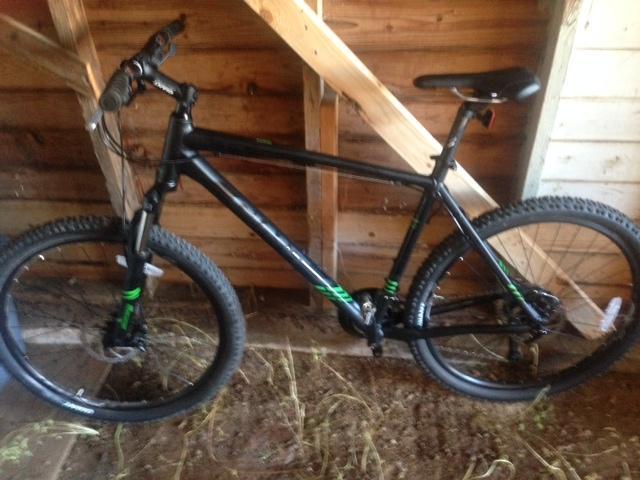 Giant Talon 27.5: A black mountain bike with green accents, positioned against a wooden structure inside a shed. The bike features thick tires and a sturdy frame, with handlebars visible in the foreground. The background shows wooden beams and dirt on the ground, indicating a rustic storage area.