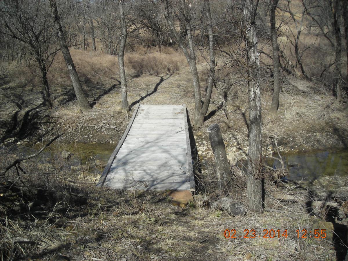 A wooden bridge spanning a small stream, surrounded by bare trees and dry grass. The setting appears to be a natural area, with a hint of early spring or late winter. Fall River State Park mountain bike trail.