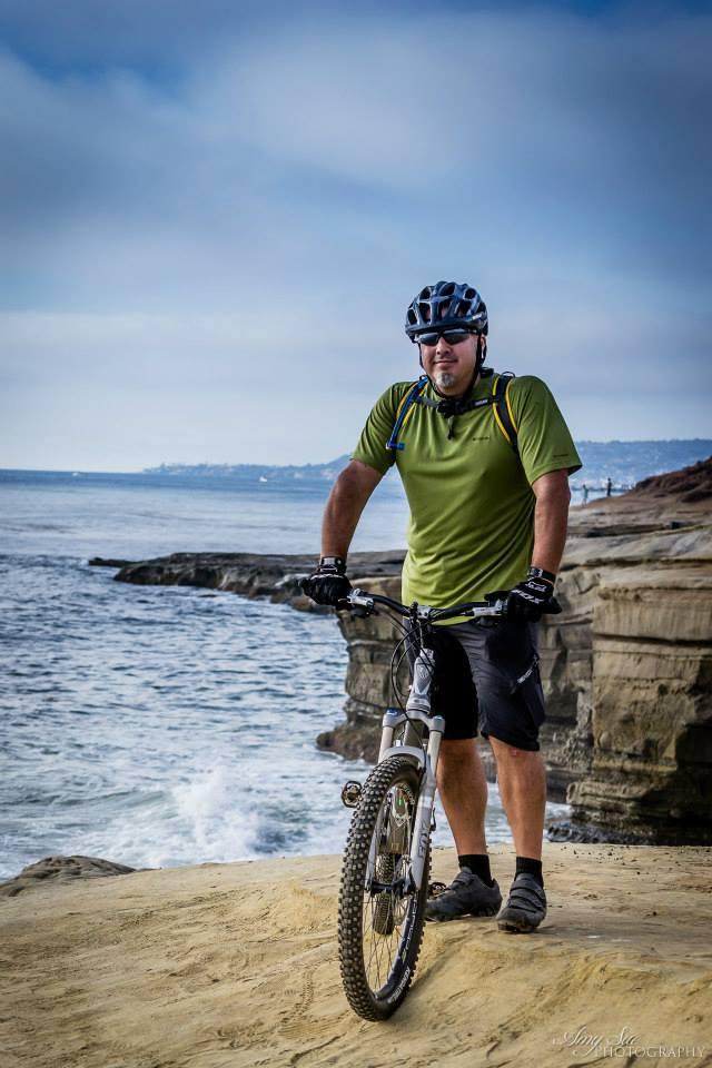 Trek Fuel EX 7: A person in a green T-shirt and black shorts stands beside a mountain bike on a sandy cliff overlooking the ocean. He is wearing a helmet and sunglasses, with a scenic view of the coastline and waves in the background under a cloudy sky.