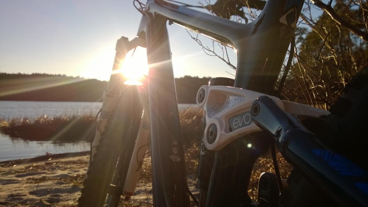 Trek Fuel EX 9.7 29: Close-up view of a mountain bike frame positioned against a serene lakeside backdrop. The sun is setting in the background, creating a warm, golden glow that highlights the bike's design and surroundings, including sandy shores and subtle vegetation.