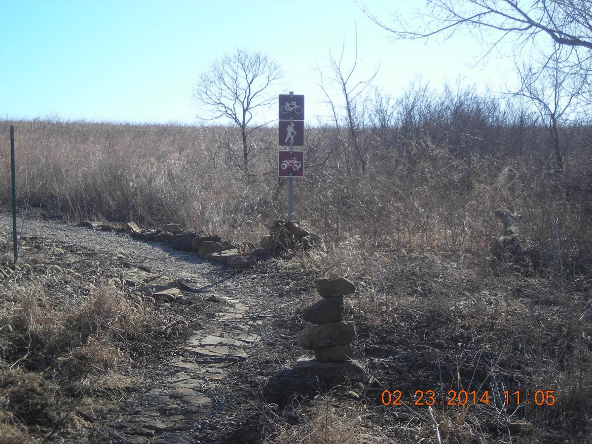 A gravel path leading into a field marked by a sign with symbols for hiking and cycling. The area is surrounded by tall, dry grass and sparse trees under a clear blue sky. Stacked rocks line the pathway's edge. Badger Creek South Trail mountain bike trail.