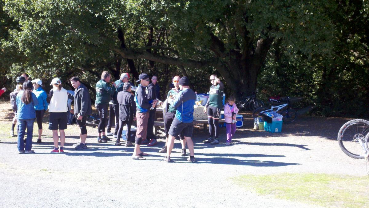 A group of people gathered outdoors near a picnic table under a large tree, engaging in conversation and enjoying refreshments. Some individuals are wearing athletic clothing and helmets, suggesting they have participated in a biking or outdoor event. Bicycles are visible in the background, along with various supplies.