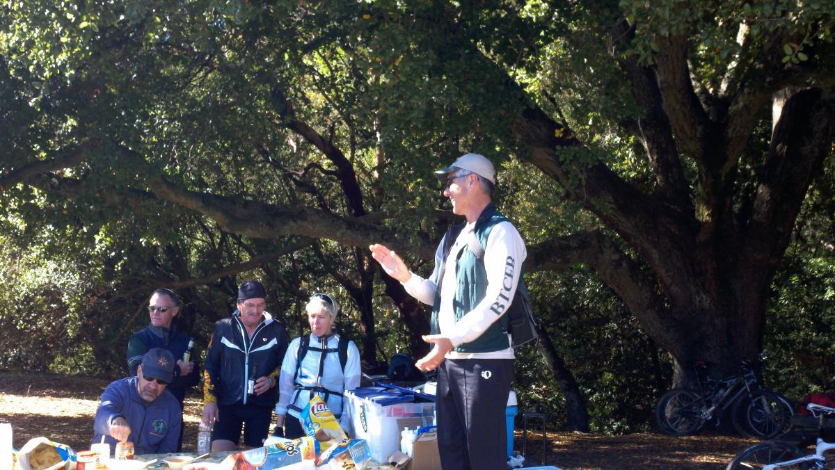 A group of people gathered outdoors under a large tree, engaged in conversation. One person stands in front of a table filled with snacks and drinks, gesturing as if explaining something. The others, wearing casual outdoor clothing, listen attentively. Bikes are parked nearby, indicating a recreational outing.