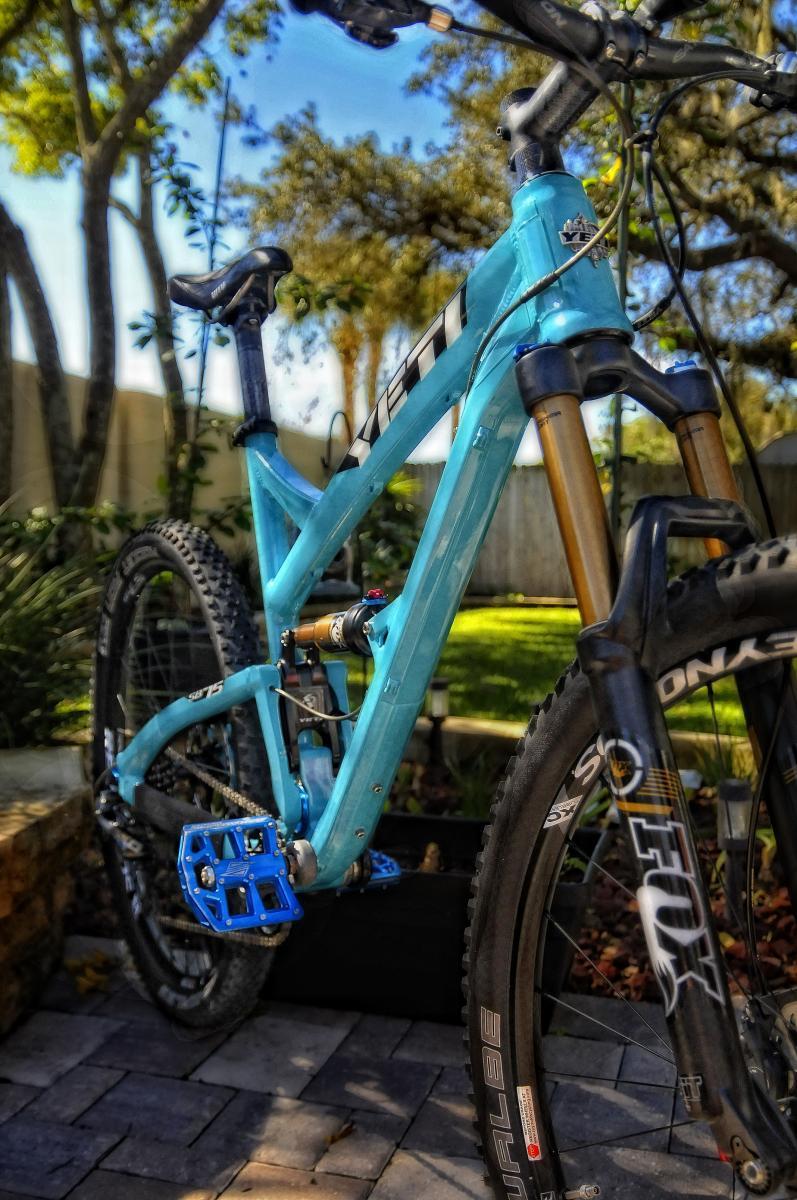Yeti SB-75: Alt text: A close-up view of a blue mountain bike parked outdoors, featuring a sturdy frame, sleek tires, and detailed components, surrounded by greenery and sunlight.
