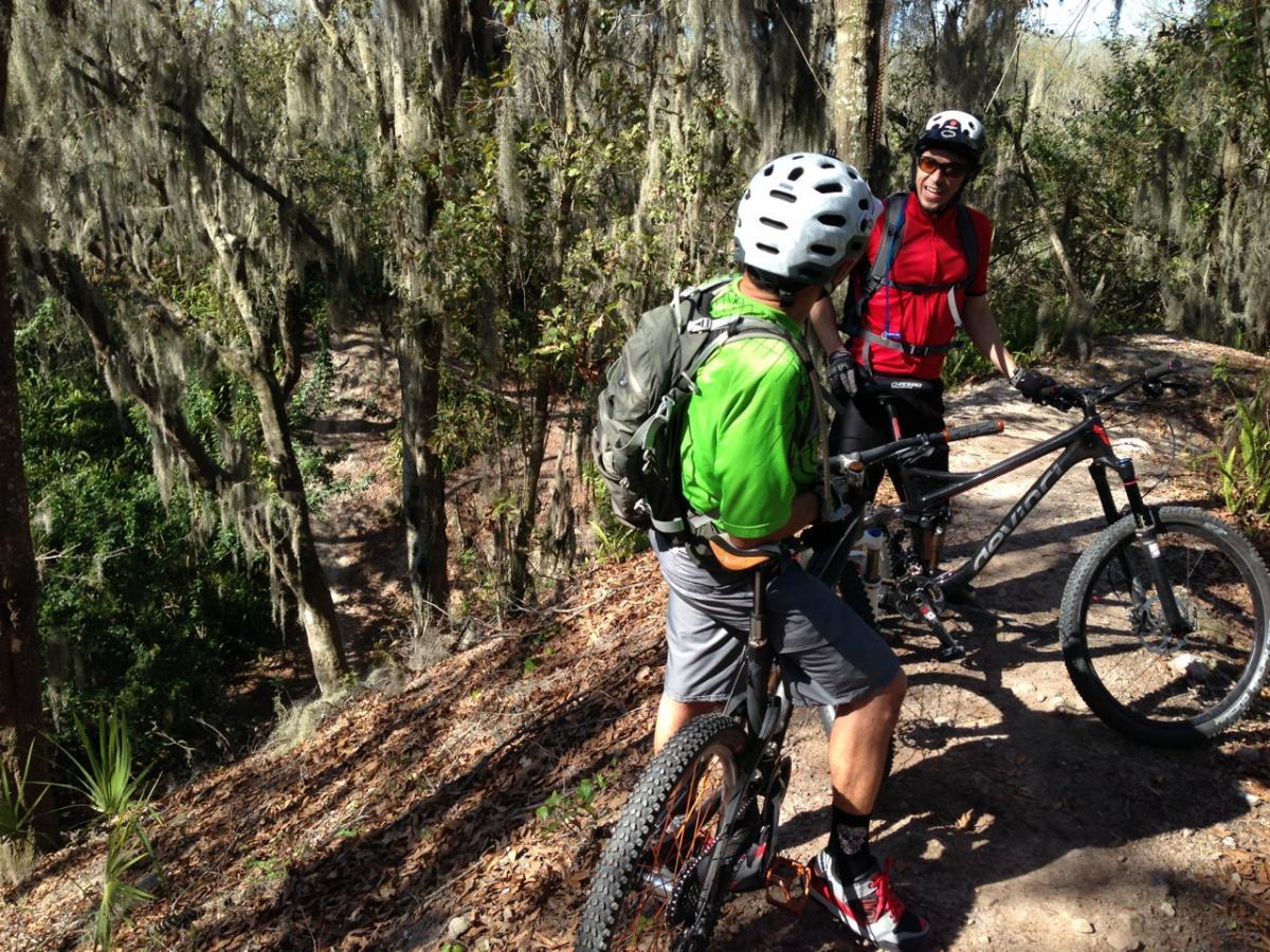 Two mountain bikers pause on a wooded trail, surrounded by tall trees draped with Spanish moss. One biker, wearing a green shirt and helmet, leans against his bike, while the other, dressed in a red shirt and black helmet, smiles and engages in conversation. The path ahead winds through the lush greenery of the forest. Alafia River State Park mountain bike trail.