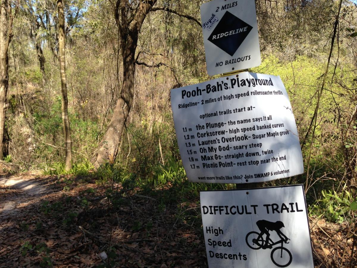 Text description of a trail sign in a wooded area, indicating the start of the Ridgeline trail, which is two miles long and noted for its high-speed rollercoaster thrills. The sign lists optional trails with names and brief descriptions, such as "the Plunge," "Corkscrew," and "Oh My God." Another sign indicates "Difficult Trail" with a warning for high-speed descents. The surroundings feature trees and natural foliage. Balm Boyette Scrub Preserve mountain bike trail.