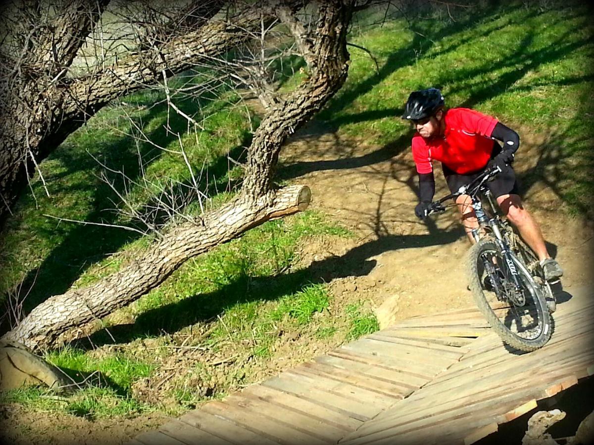 A mountain biker in a red jersey navigates a wooden trail on a sunny day, surrounded by green grass and bare trees. The biker leans into a turn, showcasing skill and balance. Lagoon Valley mountain bike trail.