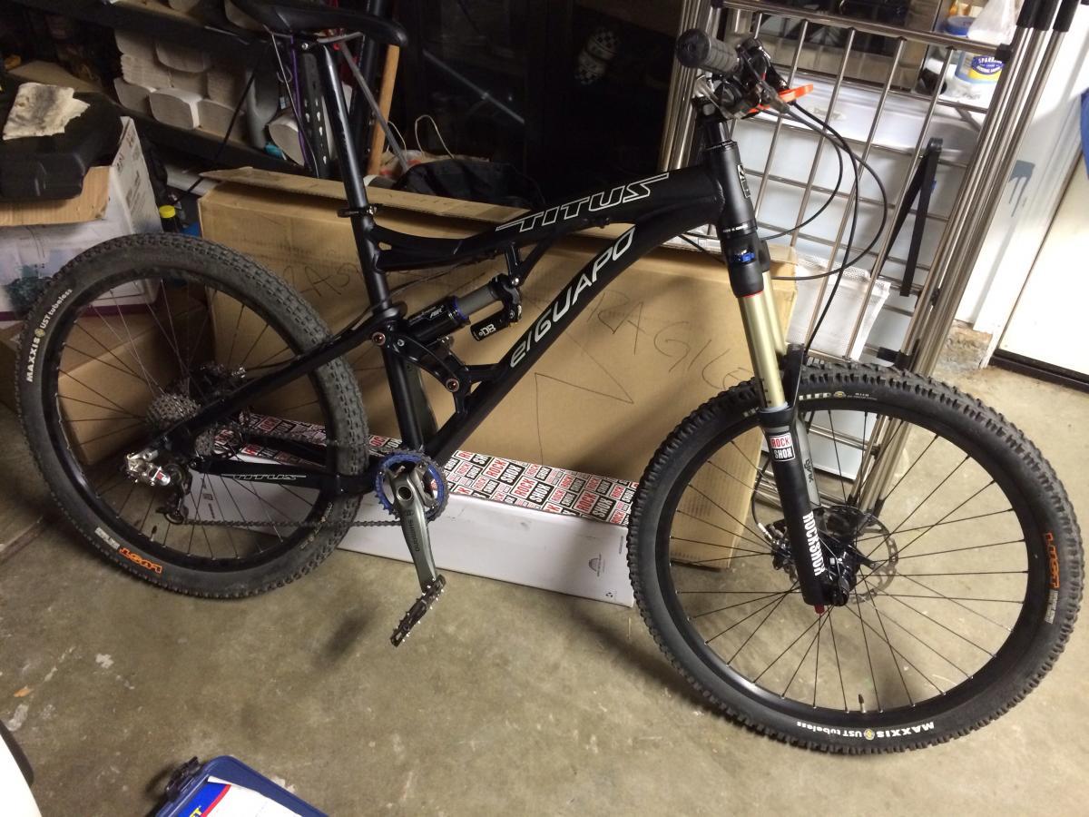 Titus El Guapo: A black mountain bike leaning against cardboard boxes in a garage. The bike features front and rear suspension, a single chainring, and wide tires designed for rough terrain. The environment is cluttered, with tools and other items visible in the background.