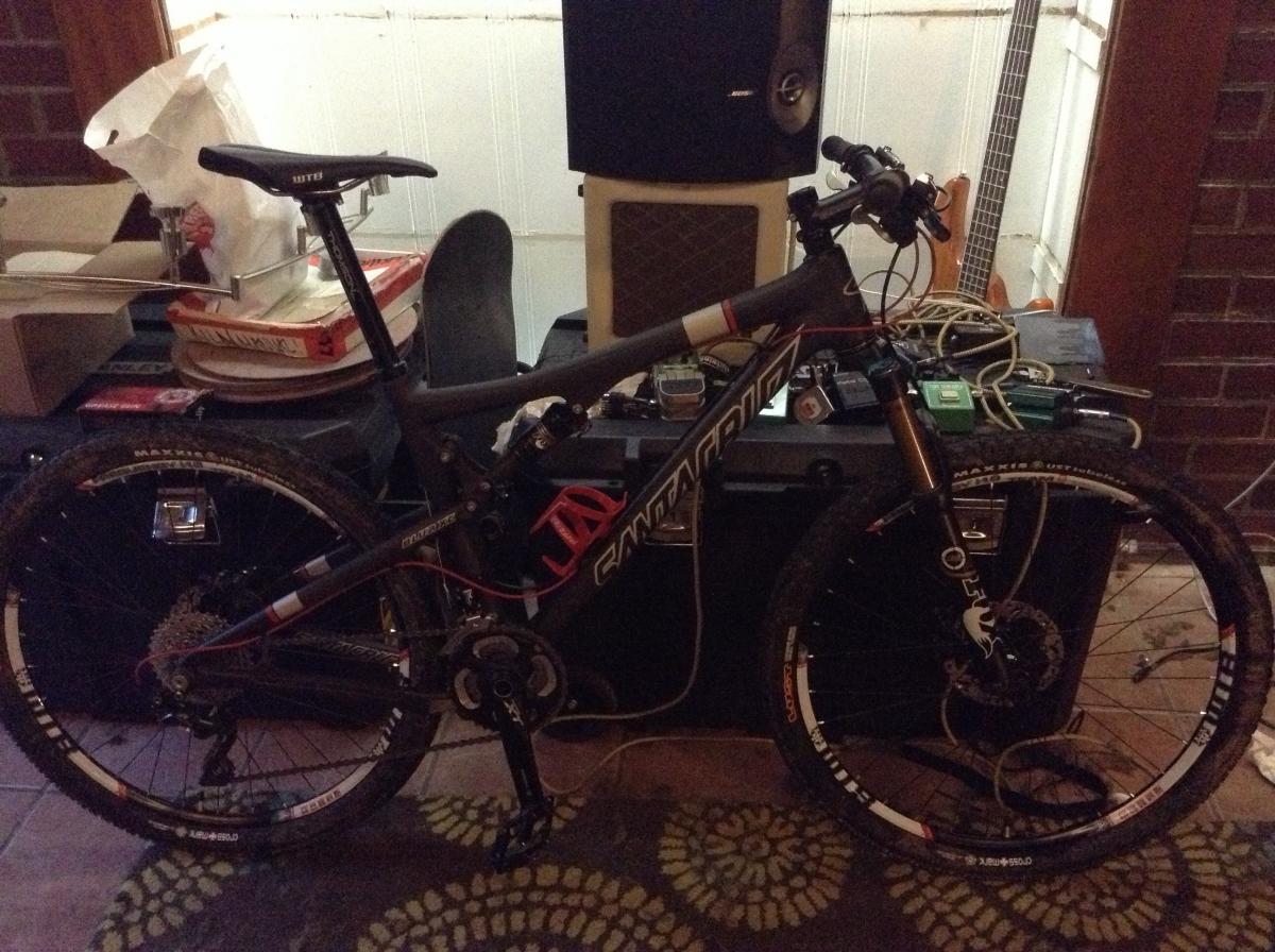 Santa Cruz Blur: A mountain bike is positioned in a cluttered indoor setting with various items in the background, including boxes and music equipment. The bike features a sleek design with a black frame, visible gears, and tires with some dirt, suggesting it has been used off-road. The surroundings are dimly lit, creating a casual atmosphere.