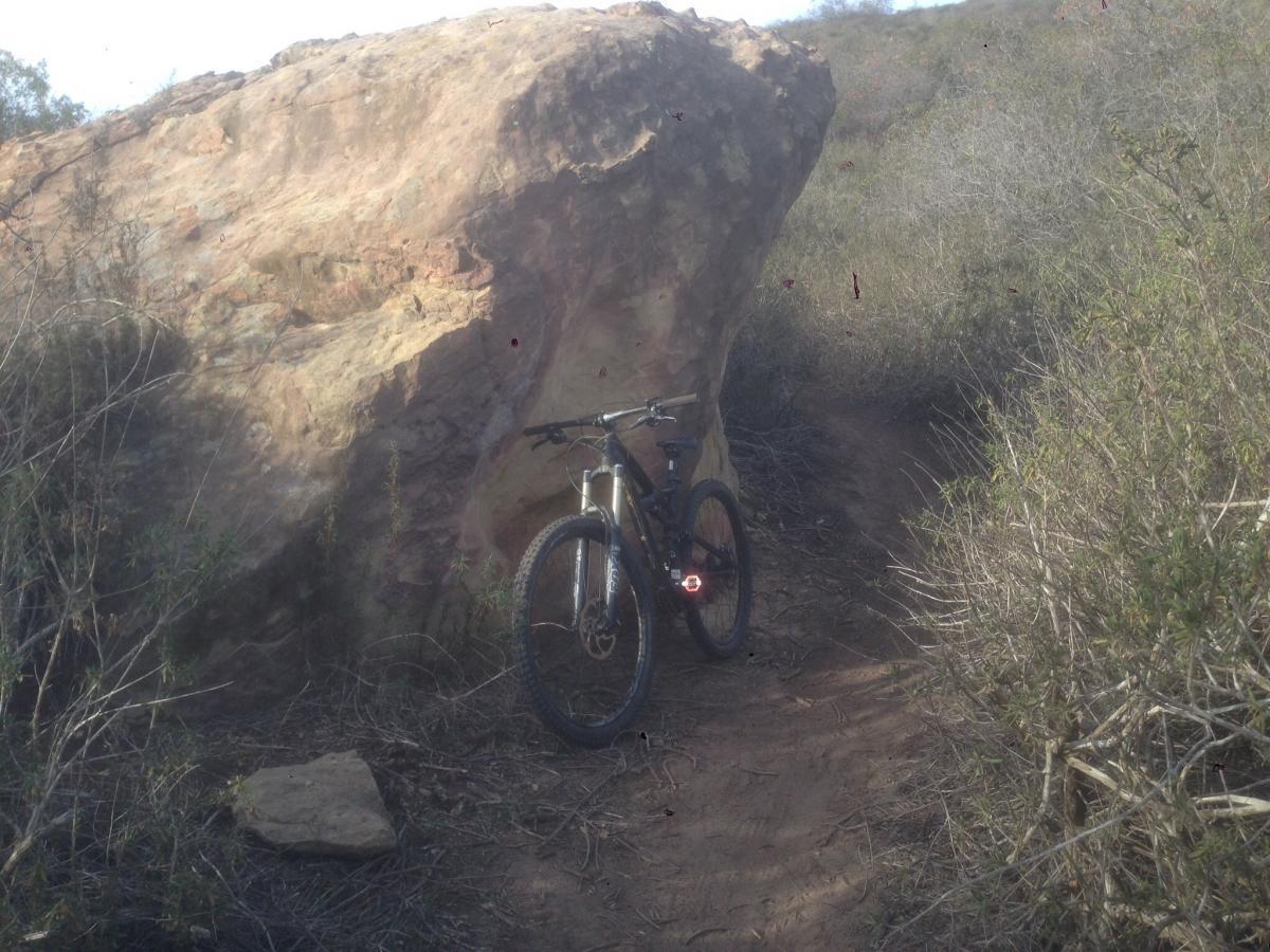 Specialized Enduro Comp 29: A mountain bike parked next to a large rock formation on a dirt trail surrounded by shrubs and vegetation. The scene captures a rocky landscape in a natural outdoor setting.
