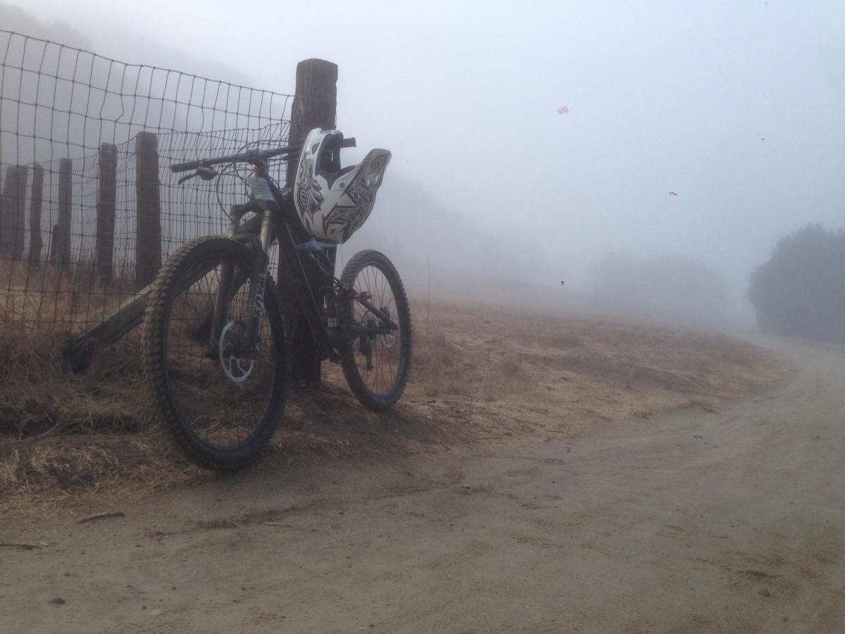 Specialized Enduro Comp 29: A mountain bike leans against a wooden fence in a foggy environment, with grassy terrain and a dirt path visible in the background. The scene is shrouded in mist, creating a tranquil and mysterious atmosphere.