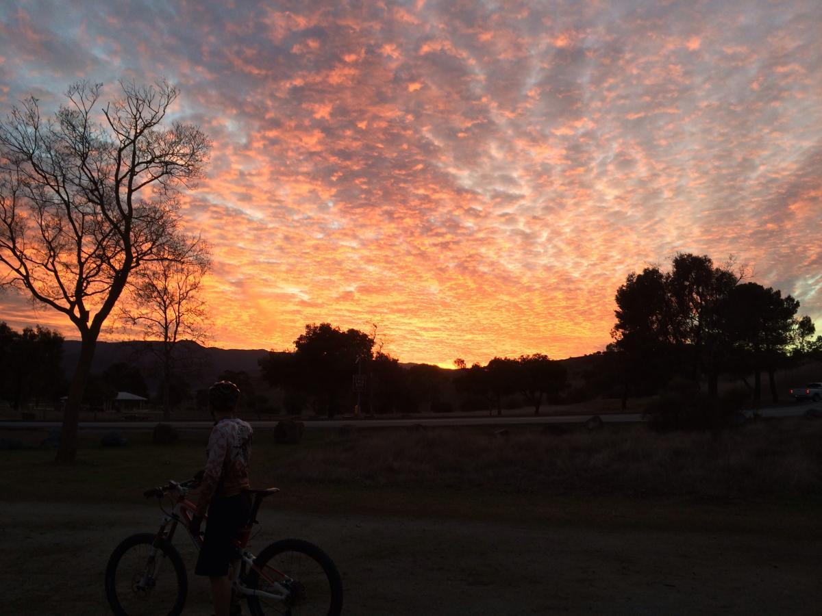 A silhouette of a person standing next to a bicycle, observing a vibrant sunset with hues of orange, pink, and purple in the sky. Leafless trees and rolling hills are visible in the background, creating a serene outdoor scene. Santa Teresa Park mountain bike trail.