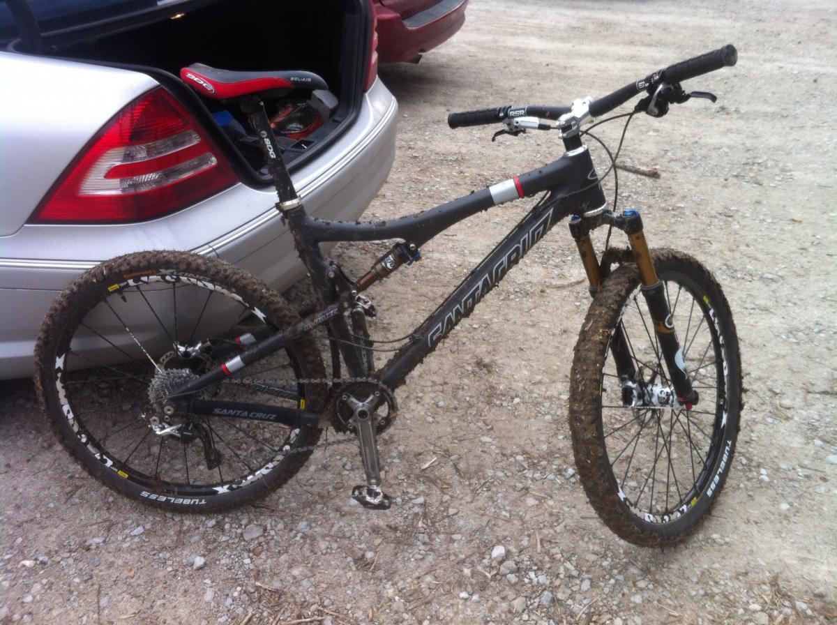 Santa Cruz Blur: A black mountain bike with muddy tires parked beside a silver car. The bike features a prominent "Santa Cruz" logo, front suspension forks, and dirt on its frame, indicating recent use on off-road trails. The car's trunk is open, revealing gear and equipment. The background shows a gravel surface.
