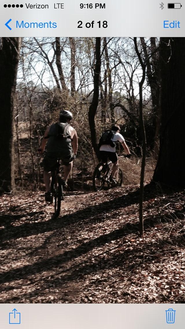 Two cyclists riding mountain bikes on a wooded trail, surrounded by trees and fallen leaves, with one cyclist wearing a helmet and a sleeveless shirt, while the other is in a white shirt and a backpack. The scene appears sunny and calm, suggesting an outdoor adventure. Spadra Creek Nature Trail mountain bike trail.