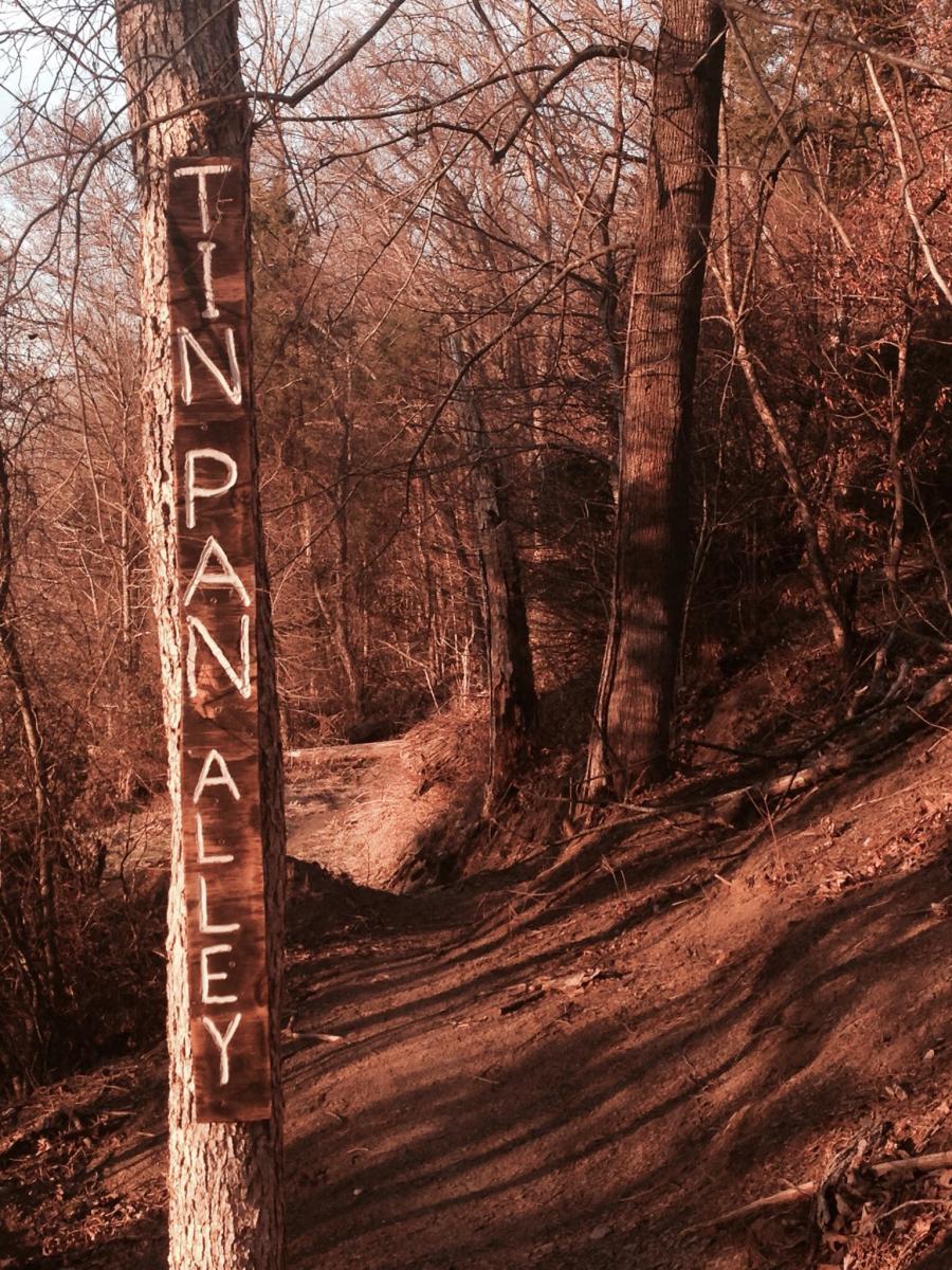 A wooden signpost labeled "TIN PAN ALLEY" is attached to a tree on a forested trail. The path curves in the background, surrounded by bare trees and earthy tones of the landscape. Spadra Creek Nature Trail mountain bike trail.