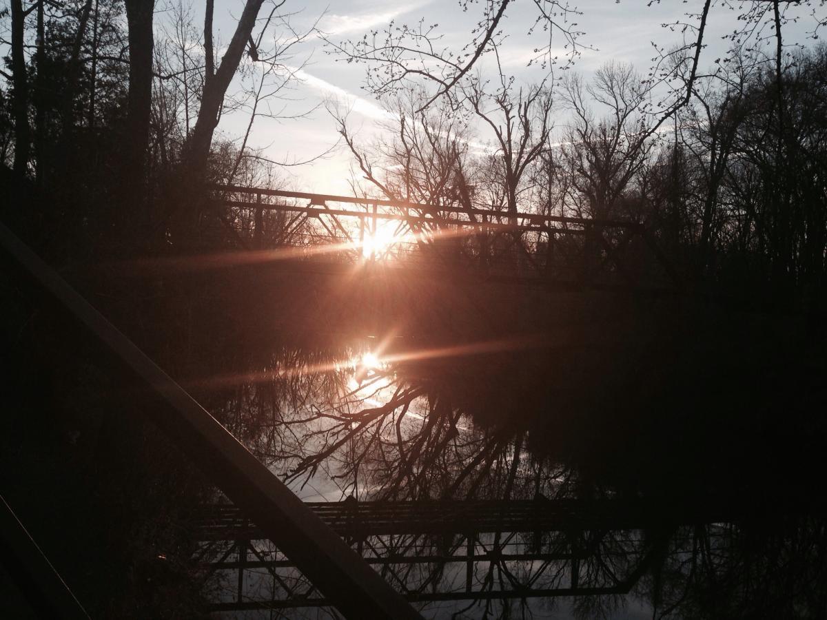 A serene landscape featuring a river at sunset, with sunlight reflecting off the water's surface. Silhouetted trees line the riverbank, and an old wooden bridge spans the river in the background. The scene captures a tranquil moment in nature, with a soft, warm glow from the setting sun. Spadra Creek Nature Trail mountain bike trail.
