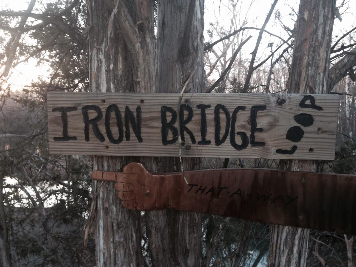 Signpost indicating "Iron Bridge" with an arrow pointing to the right. The sign is made of wood, featuring black letters and a carved wooden hand pointing towards the direction. In the background, trees are visible along with a glimpse of water. Spadra Creek Nature Trail mountain bike trail.