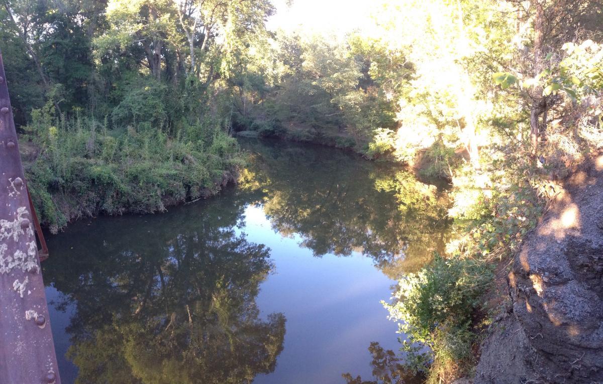 A serene view of a calm river surrounded by lush greenery, with reflections of trees on the water's surface. Sunlight filters through the leaves, creating a peaceful and tranquil atmosphere. Spadra Creek Nature Trail mountain bike trail.