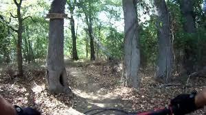 A dirt trail winding through a wooded area, flanked by tall trees. A sign hangs on one of the trees, indicating the direction of the trail, while fallen leaves cover the ground. The scene suggests a peaceful environment for cycling or hiking. Spadra Creek Nature Trail mountain bike trail.