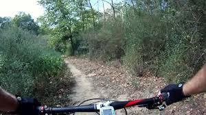 Mountain bike rider's perspective on a trail surrounded by trees and shrubs, with a path winding ahead. A portion of the bike handlebars and hands wearing gloves are visible in the foreground. Spadra Creek Nature Trail mountain bike trail.