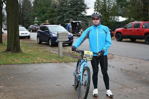 Surly Karate Monkey: A cyclist in a blue jacket and black pants stands beside a mountain bike, wearing a helmet and race number on their chest. In the background, cars are parked along a tree-lined street, with a mailbox visible. The scene is set on a cloudy day with scattered leaves on the ground.