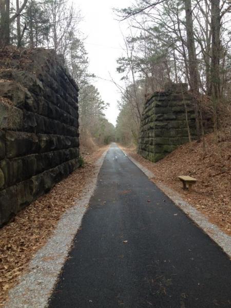 A narrow, paved pathway stretches between two large, weathered stone pillars, surrounded by trees and fallen leaves. The scene is somewhat isolated, evoking a feeling of tranquility and nature. A wooden bench sits to the side of the trail, suggesting a space for resting and enjoying the surrounding landscape. Chief Ladiga mountain bike trail.