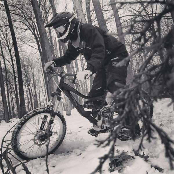 A person in a black motocross outfit and helmet rides a mountain bike through a snowy forest, surrounded by trees. The image is in black and white, emphasizing the winter environment and the dynamic motion of the rider navigating the snowy terrain. Brock Escarpment mountain bike trail.
