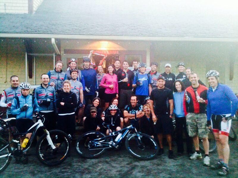A large group of cyclists gathered outside a building, smiling and posing for the camera. They are wearing a mix of cycling gear and casual clothing, with bicycles leaning against the wall. The scene is lively, with some participants holding drinks and others showing excitement. The setting appears to be an event or gathering for cycling enthusiasts.