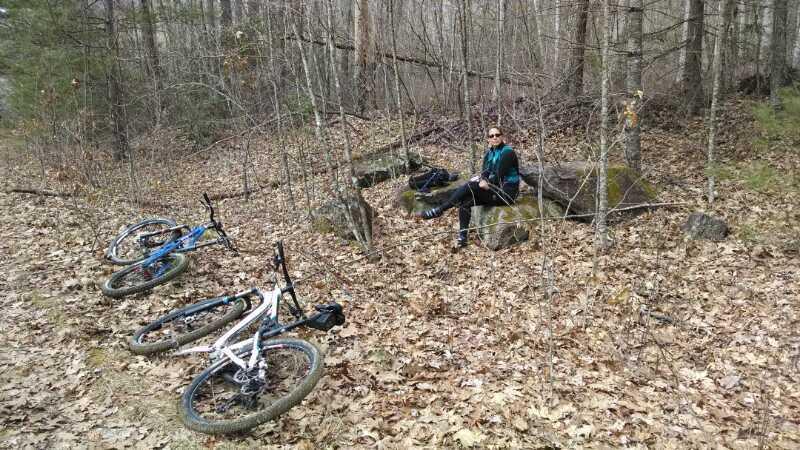 A person sitting on a rock in a forested area surrounded by trees and fallen leaves, with two mountain bikes resting nearby on the ground. The scene conveys a peaceful outdoor setting, suitable for a break during a biking adventure. North Road #453 mountain bike trail.