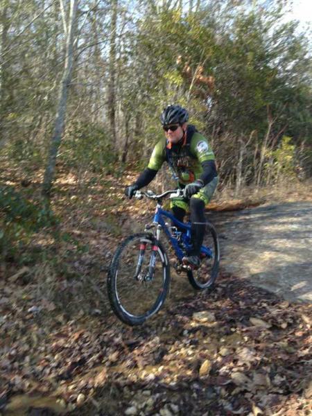 A mountain biker wearing a green and black jersey and sunglasses rides a blue bicycle along a dirt path surrounded by trees and fallen leaves. Powerline Road #451 mountain bike trail.